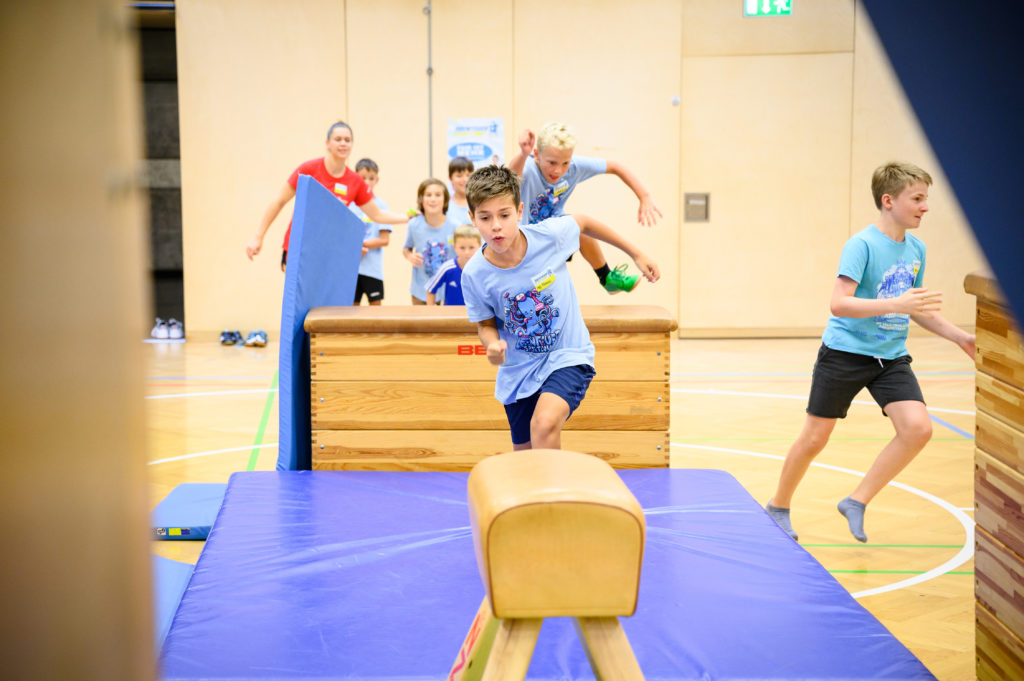 Kinder in Abenteuer-Sportcamp-Shirt machen Sport in der Halle.