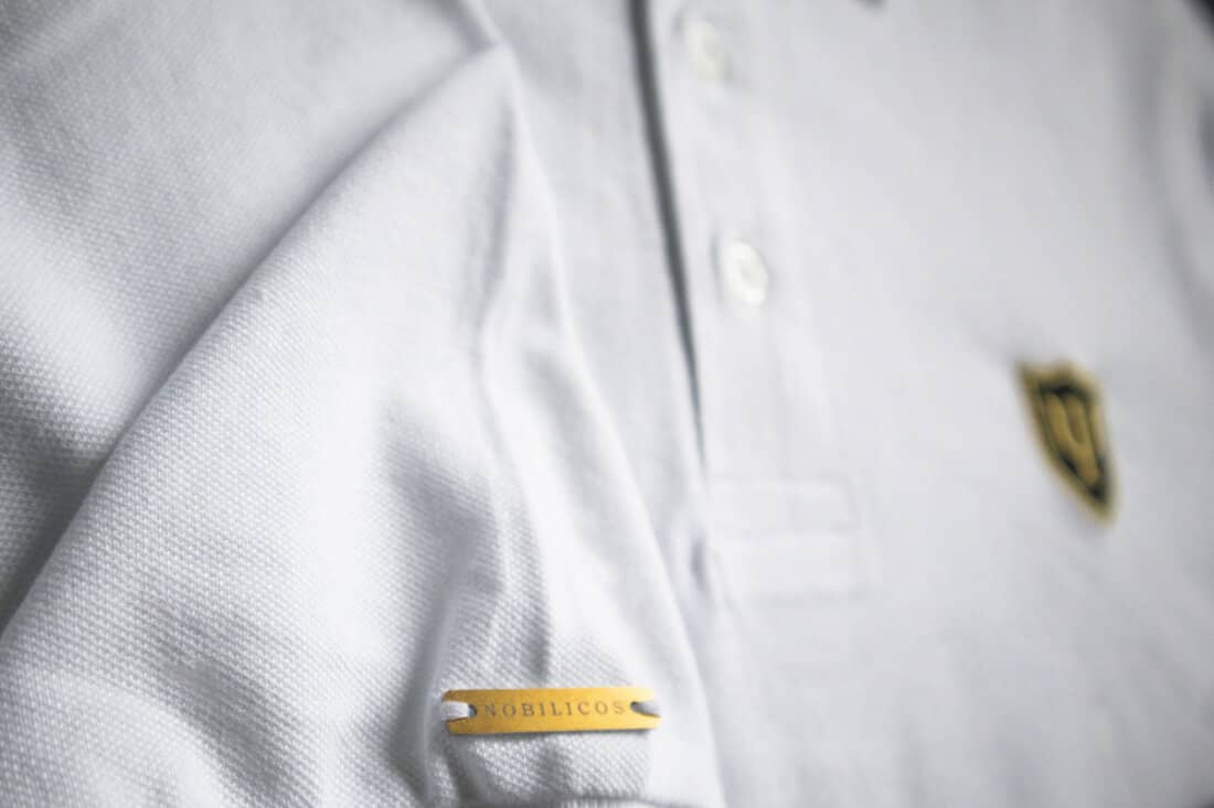Ein weißes Poloshirt mit einem goldenen, gelochten Metall-Label am Ärmel